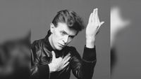 Black and white photo of a person in a leather jacket with one hand on their chest and the other raised beside their face