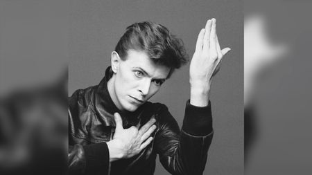 Black and white photo of a person in a leather jacket with one hand on their chest and the other raised beside their face