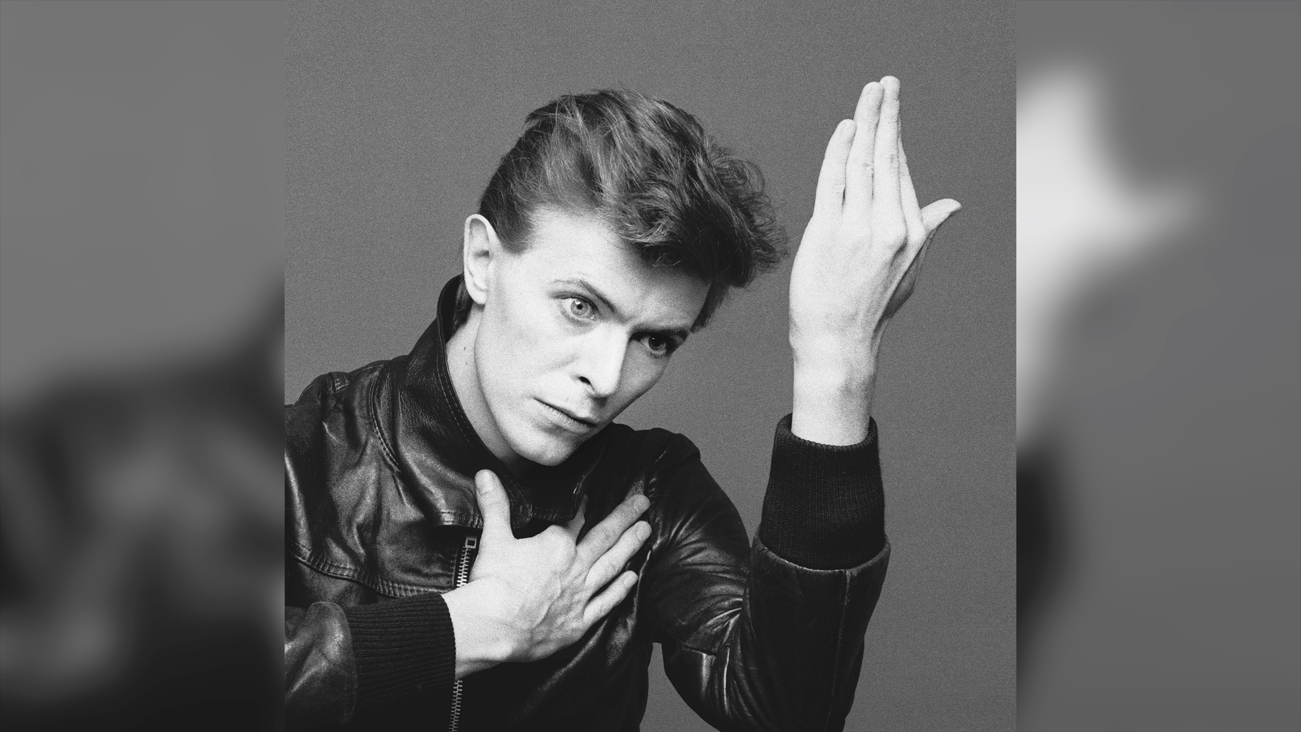 Black and white photo of a person in a leather jacket with one hand on their chest and the other raised beside their face