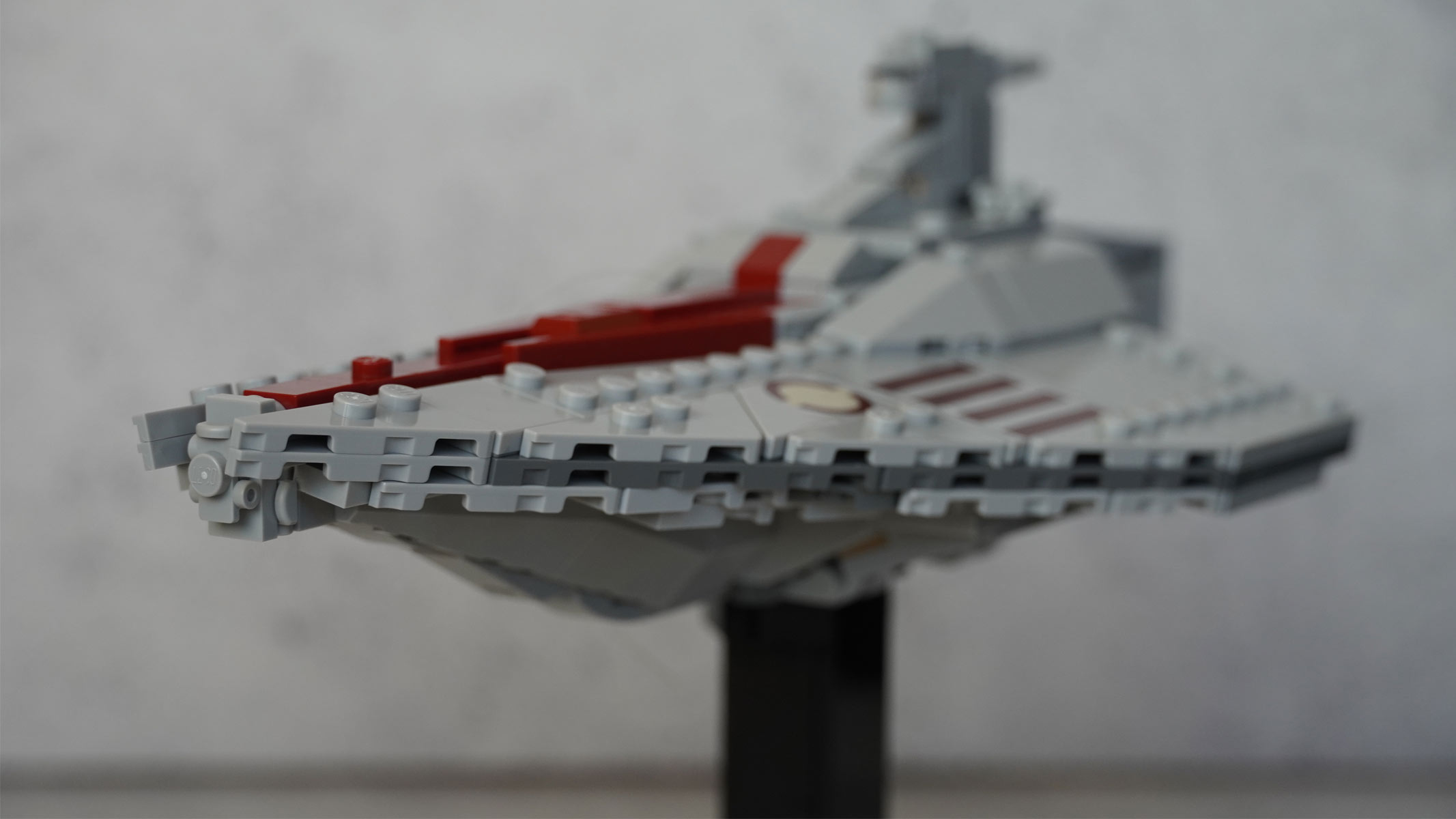 A miniature-sized long grey ship made of bricks with a red stripe running down the middle is placed on a black stand in front of a grey background.