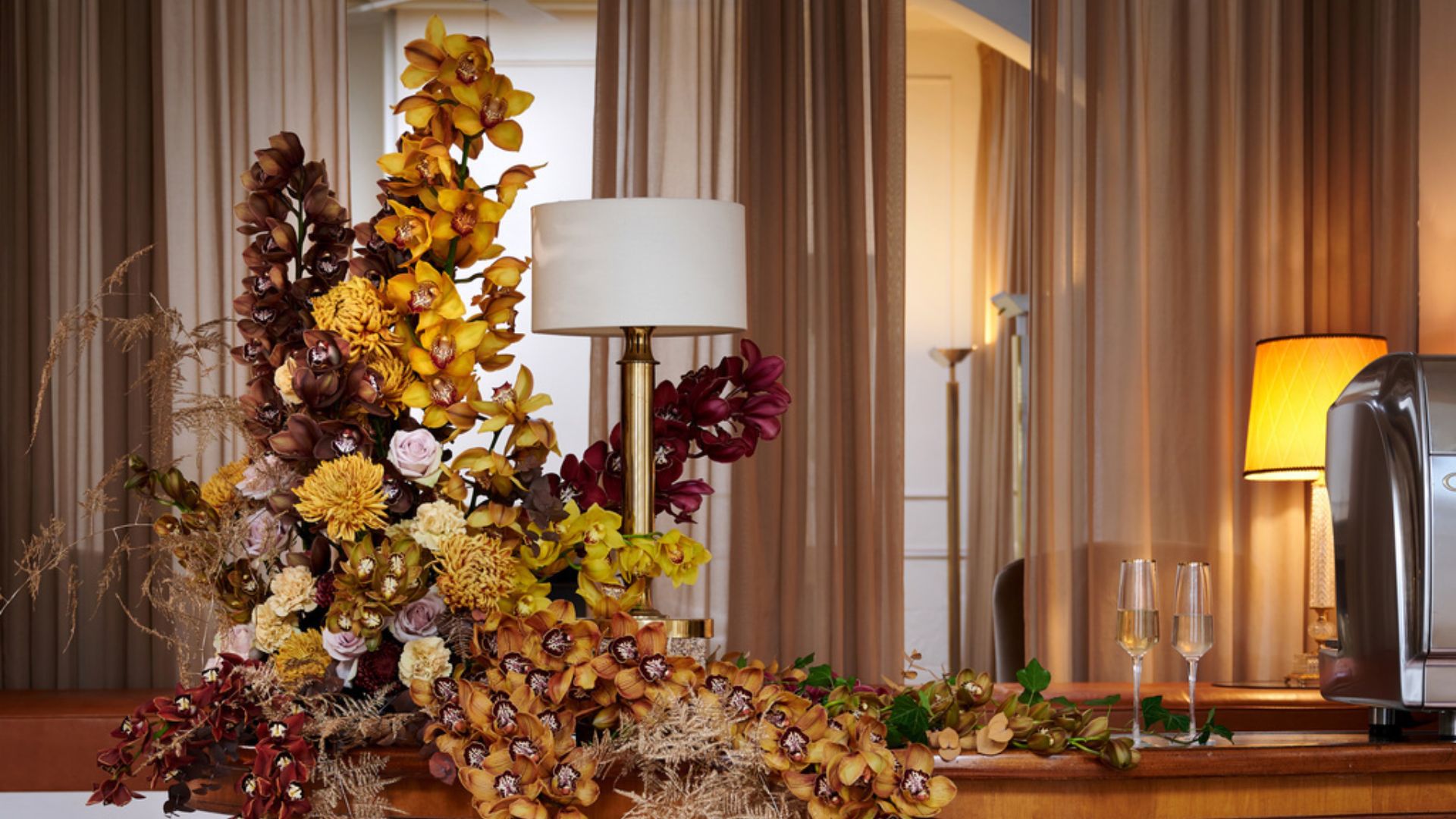 A collection of orchids displayed on a table, beside a lamp and a pair of champagne flutes