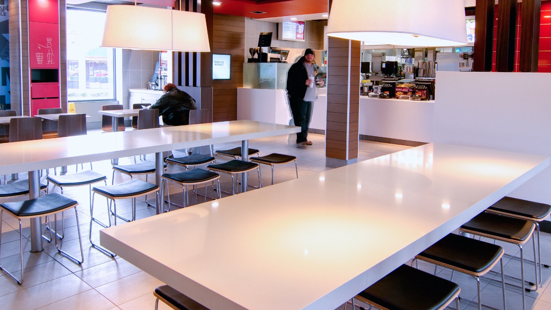 The interior of a contemporary McDonald's restaurant in Richardson, Texas, showing large white laminate tables, black metal chairs and muted wood-effect panelling under oversized white pendant lights.