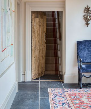 Traditional interior space with slate stone flooring, cream painted walls with artwork and traditional metal candelabra, looking onto an open rustic wooden door with carpeted stairway, blue velvet occasional chair and multi-colored patterned rug