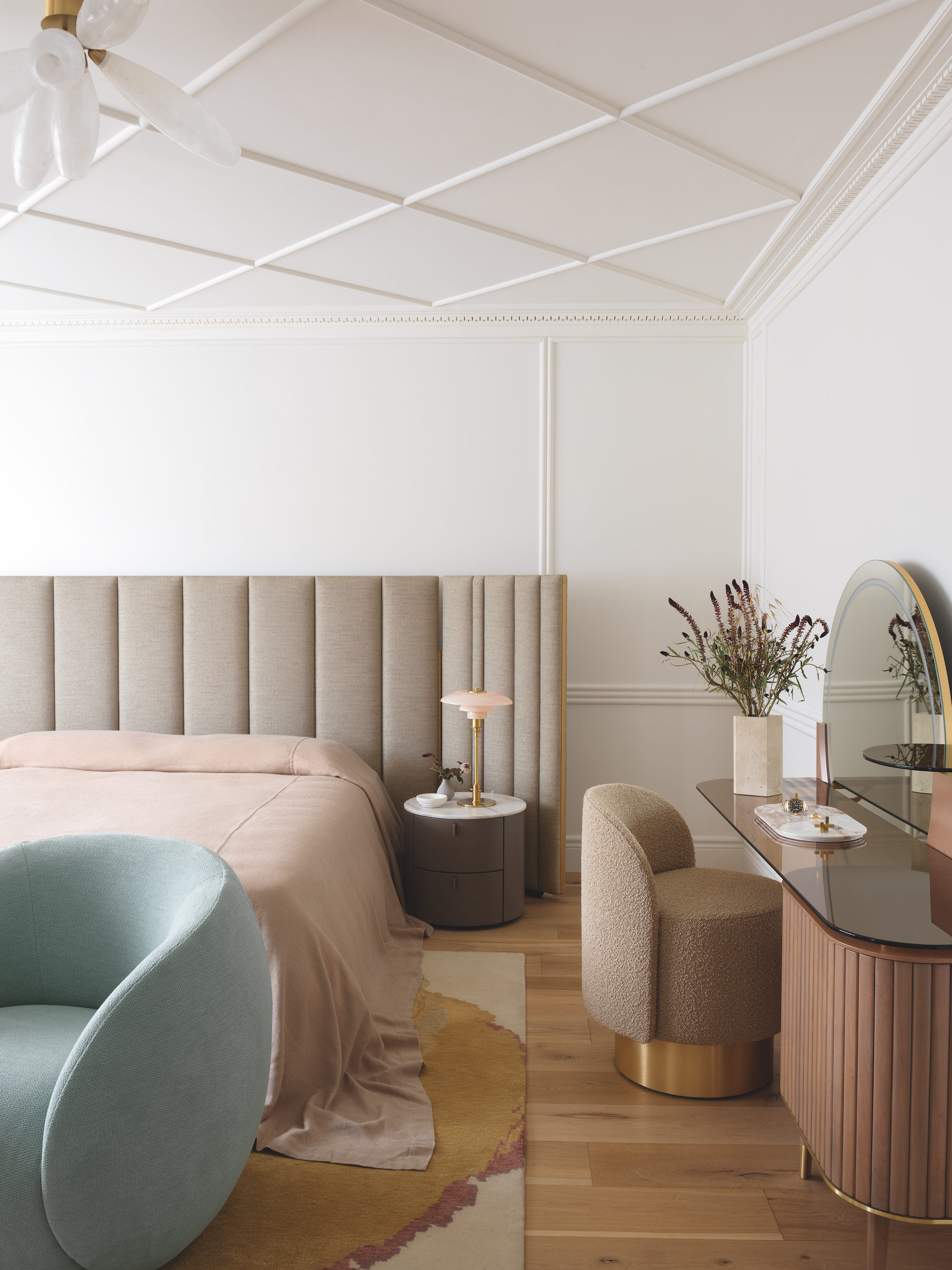 A white walled bedroom with neutral tones furniture, a blue armchair at the foot of the bed. There is a dressing table to the right and a small nightstand next to the bed, with uniquely plastered ceilings.