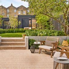 a large back garden with a black time framed studio at the end, trees and lawn in the middle and a brick patio in the foreground with a seating area