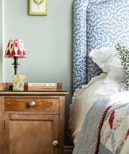 Close-up of wooden beside table with patterned lamp, books and glass. Blue and white upholstered headboatd with white bedding, throw blanket and pillow.