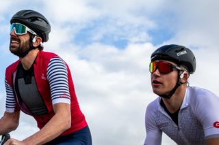 Riding with headphones. This image shows the upper body and heads of two cyclists side by side. Both have black helmets, sunglasses and a pair of Shockz headphones on. The one on the right is wearing a white short sleeve top, the on on the left has a red, white and blue short sleeved cycling top on.