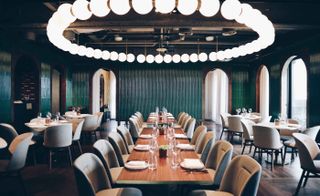 Hotel dining room with teal fluted wall tiles, long wooden tables with pale grey chairs and circle of lights hanging from ceiling