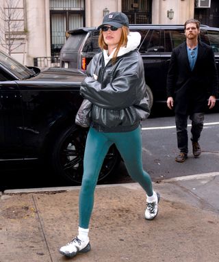 Hailey Bieber wears a black Fila hat, white hoodie, black leather jacket, emerald green leggings, white socks, and white sneakers.