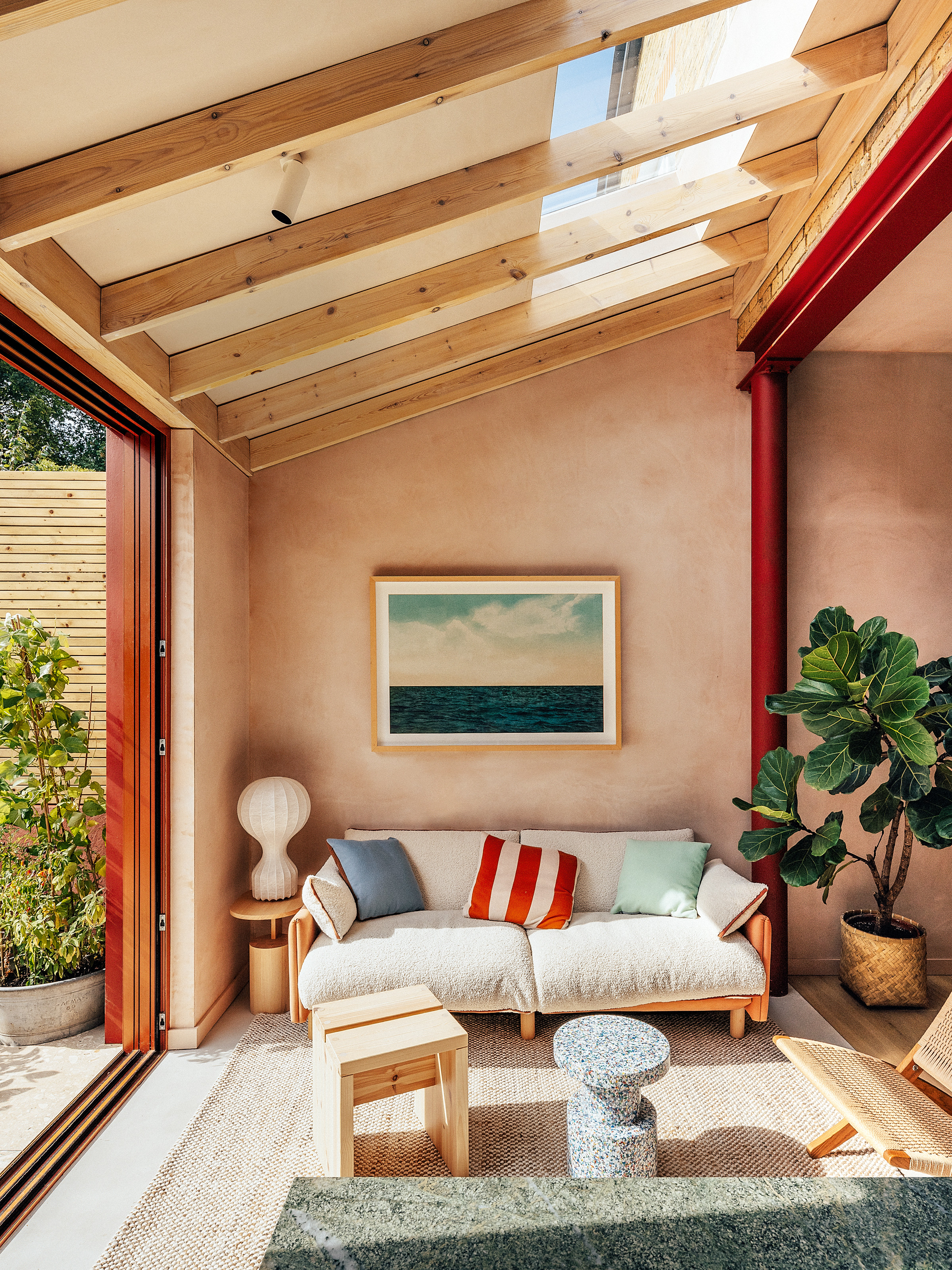 Seating area of the kitchen with plaster pink walls, red exposed structural steel, white and tan sofa, wood side tables and blue speckled stool