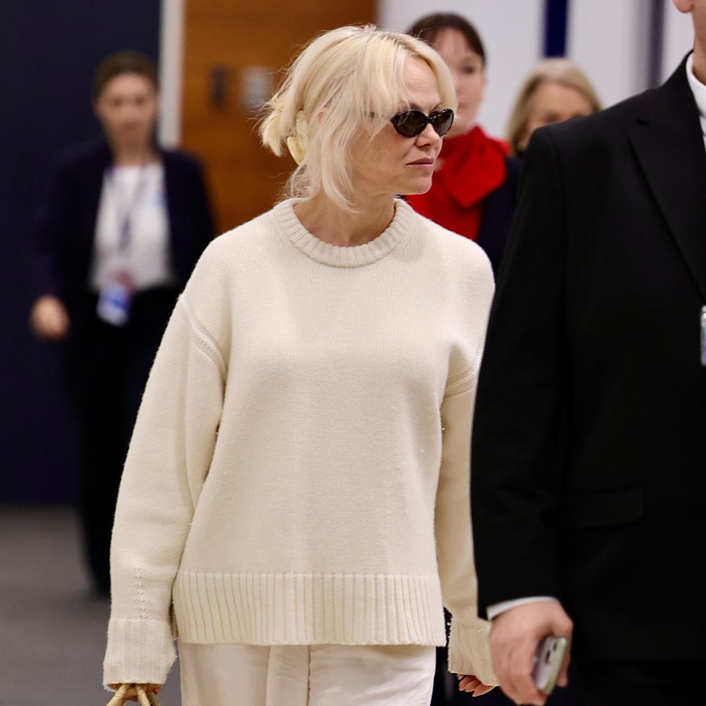 Pamela Anderson walks through the airport wearing white trainers with a wicker bag, a white jumper and white trousers.