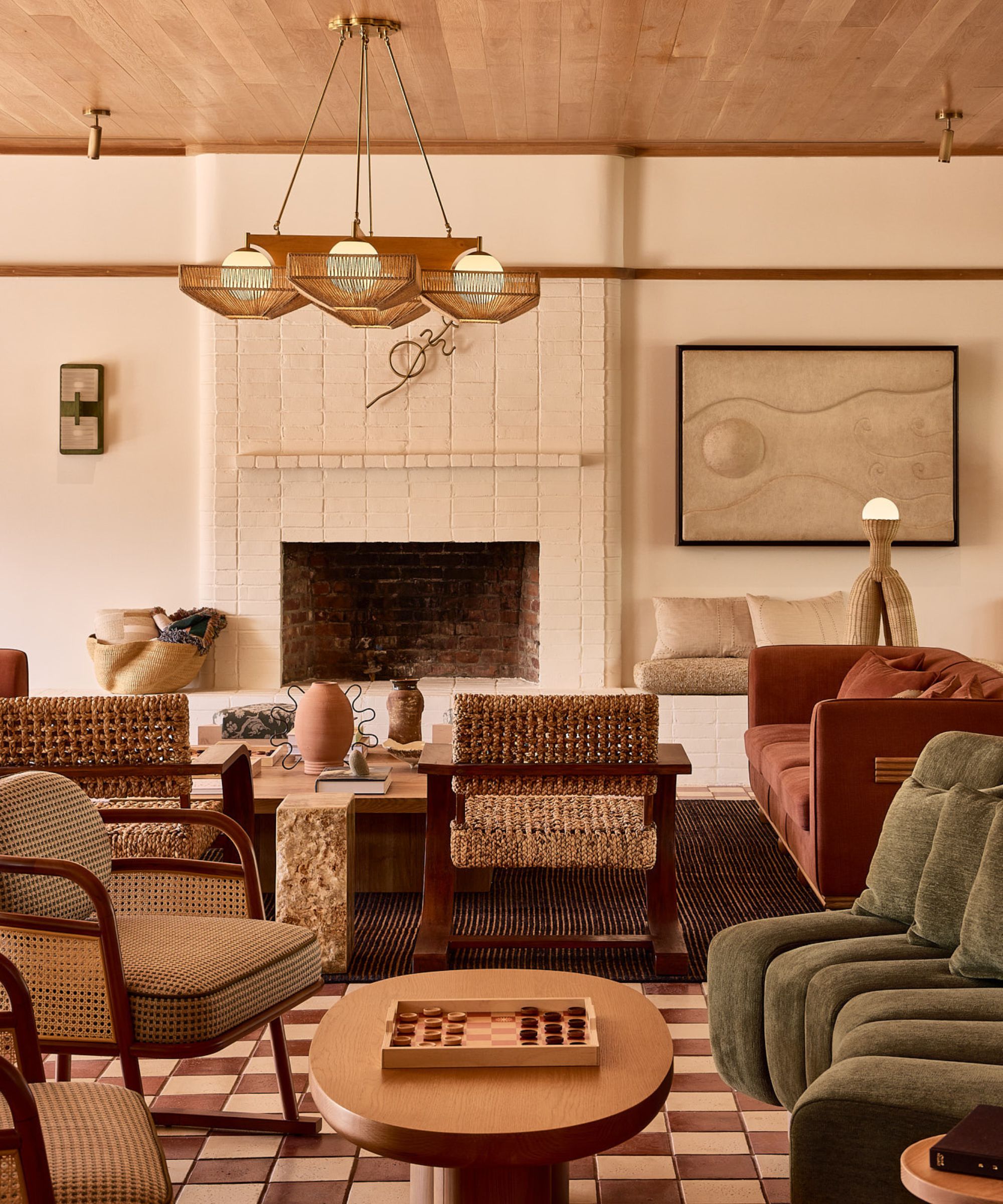 A spacious, earth-toned lounge with terracotta floor tiles, woven wicker chairs, a white brick fireplace, and a large wood-paneled ceiling