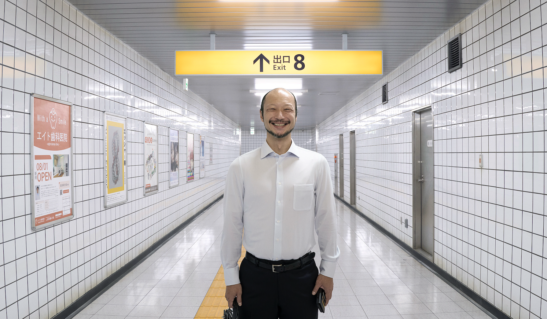 Yamato Kochi as The Walking Man, standing wearing a white shirt, black trousers and grinning at the viewer. He is standing in a subway tunnel.