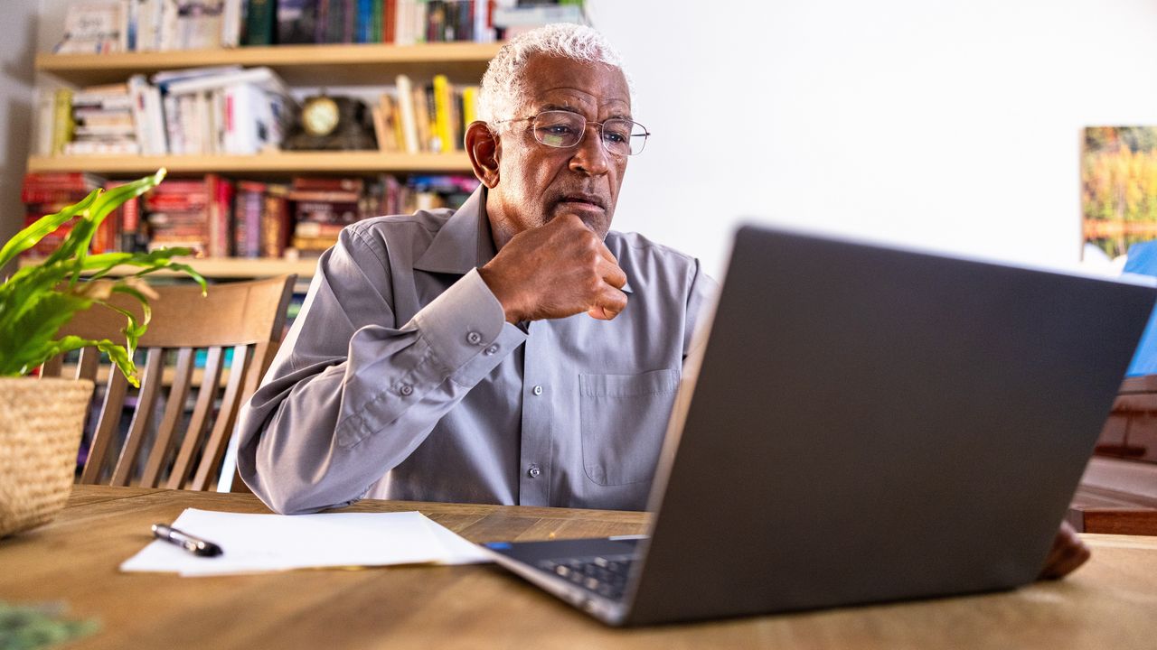 An older man looks thoughtful as he reads what&#039;s on his laptop screen at his dining room table.