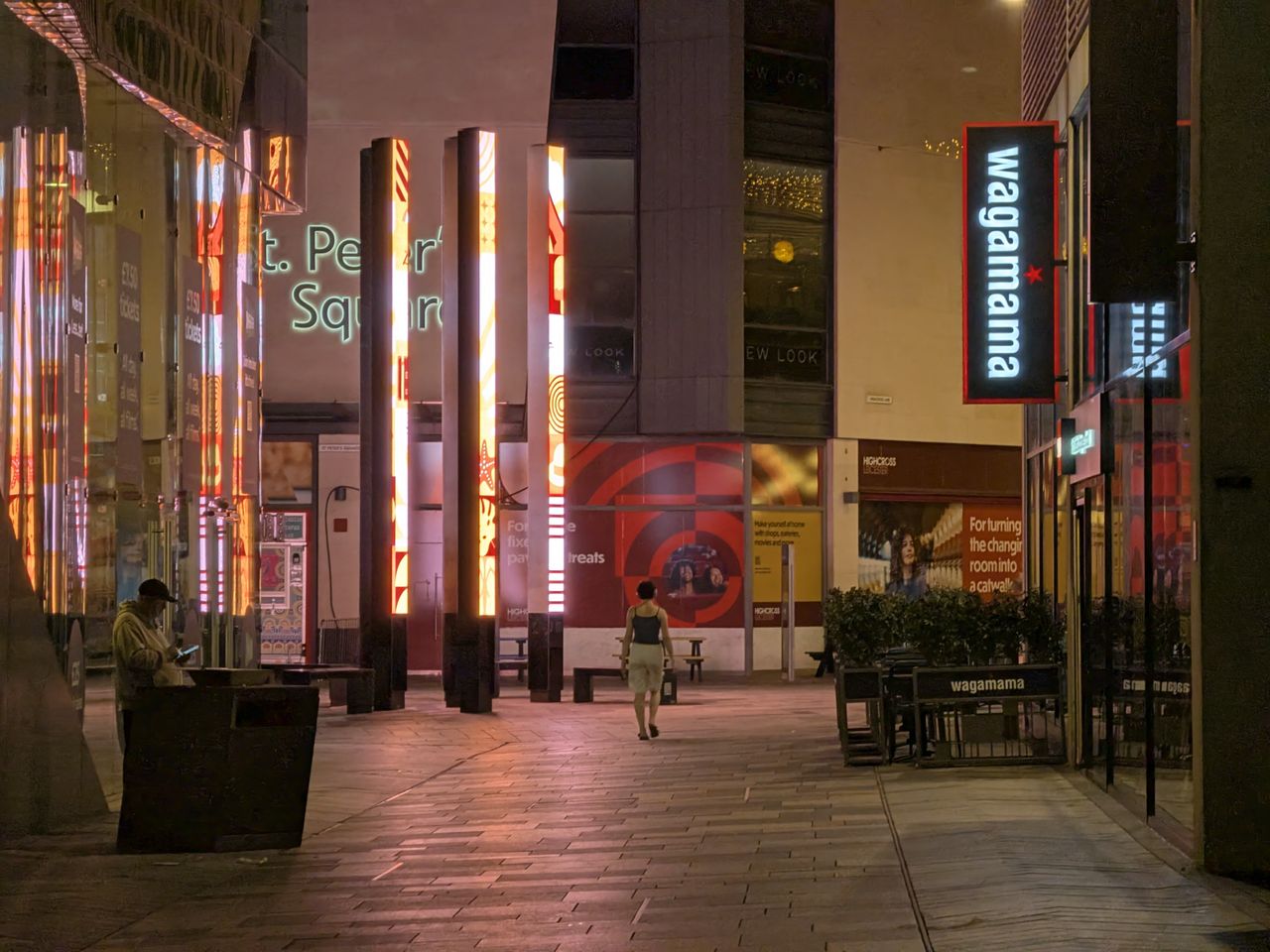 Modern pedestrian shopping street at night, with illuminated glass storefronts and reflective architecture overhead.