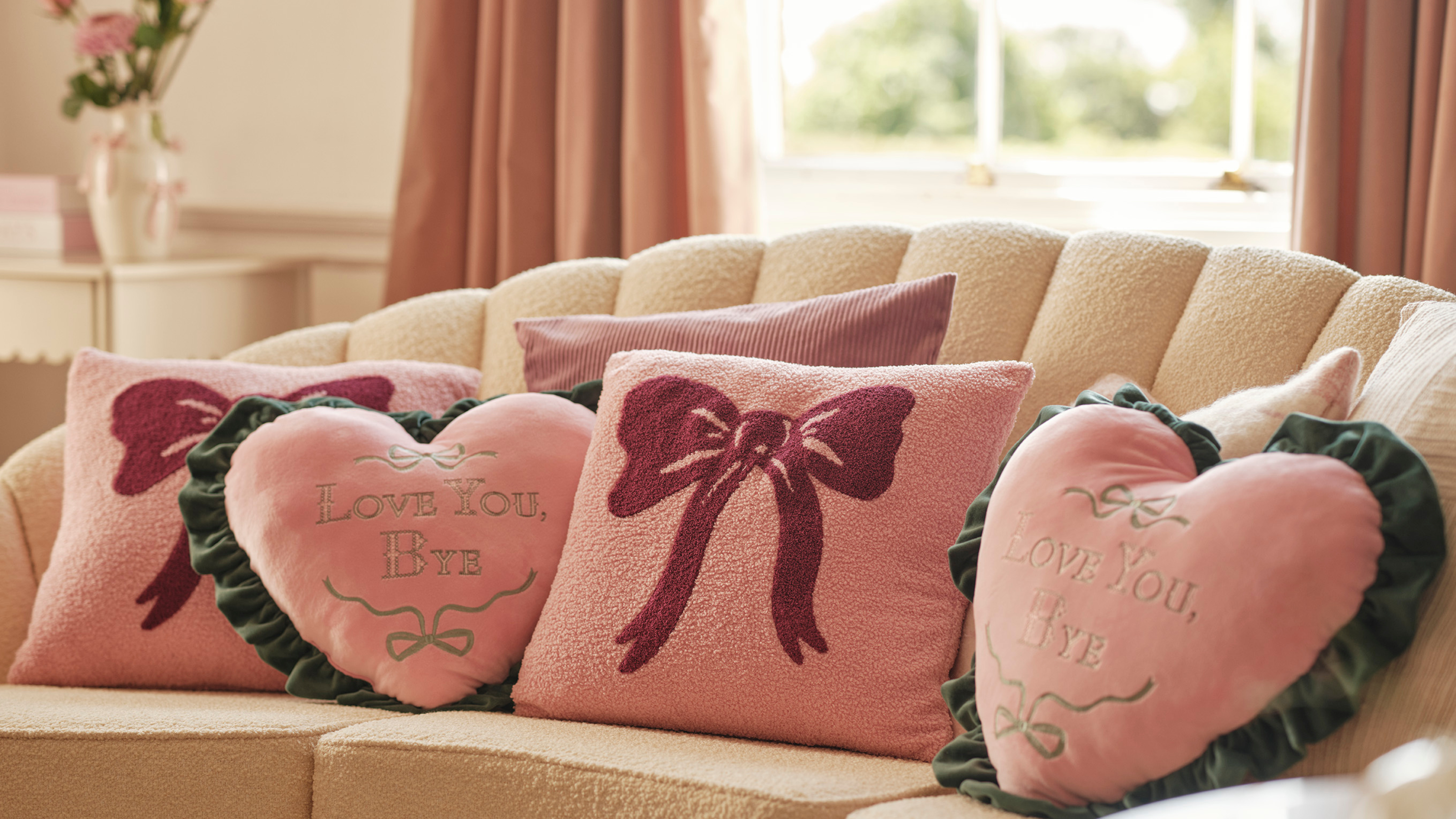 Two pink french heart cushions on a white boucle sofa. There is a pink bow cushion in the middle.