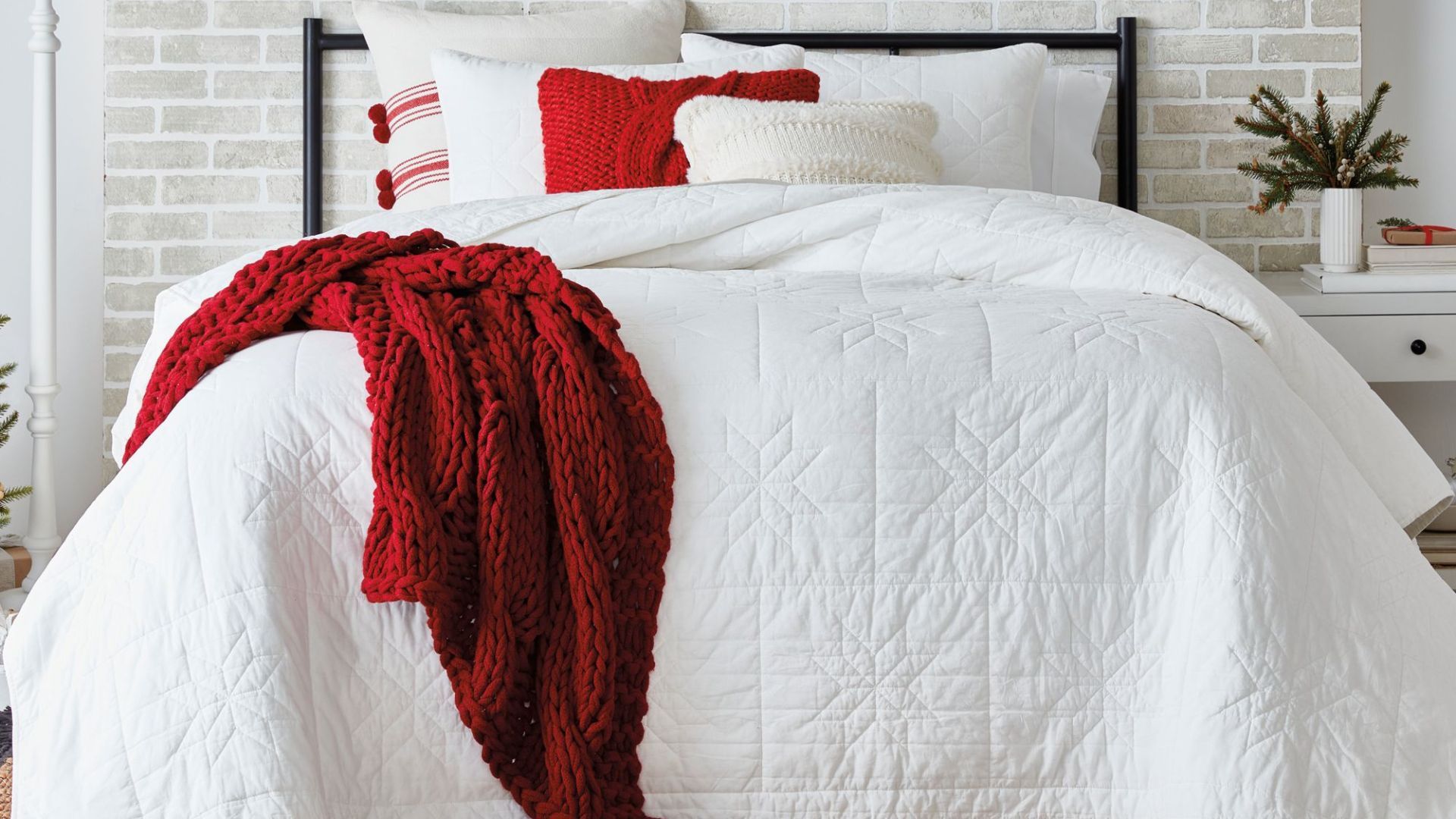A white quilt Christmas bedding set with snowflake stitching with a red knitted throw laying on the bed.