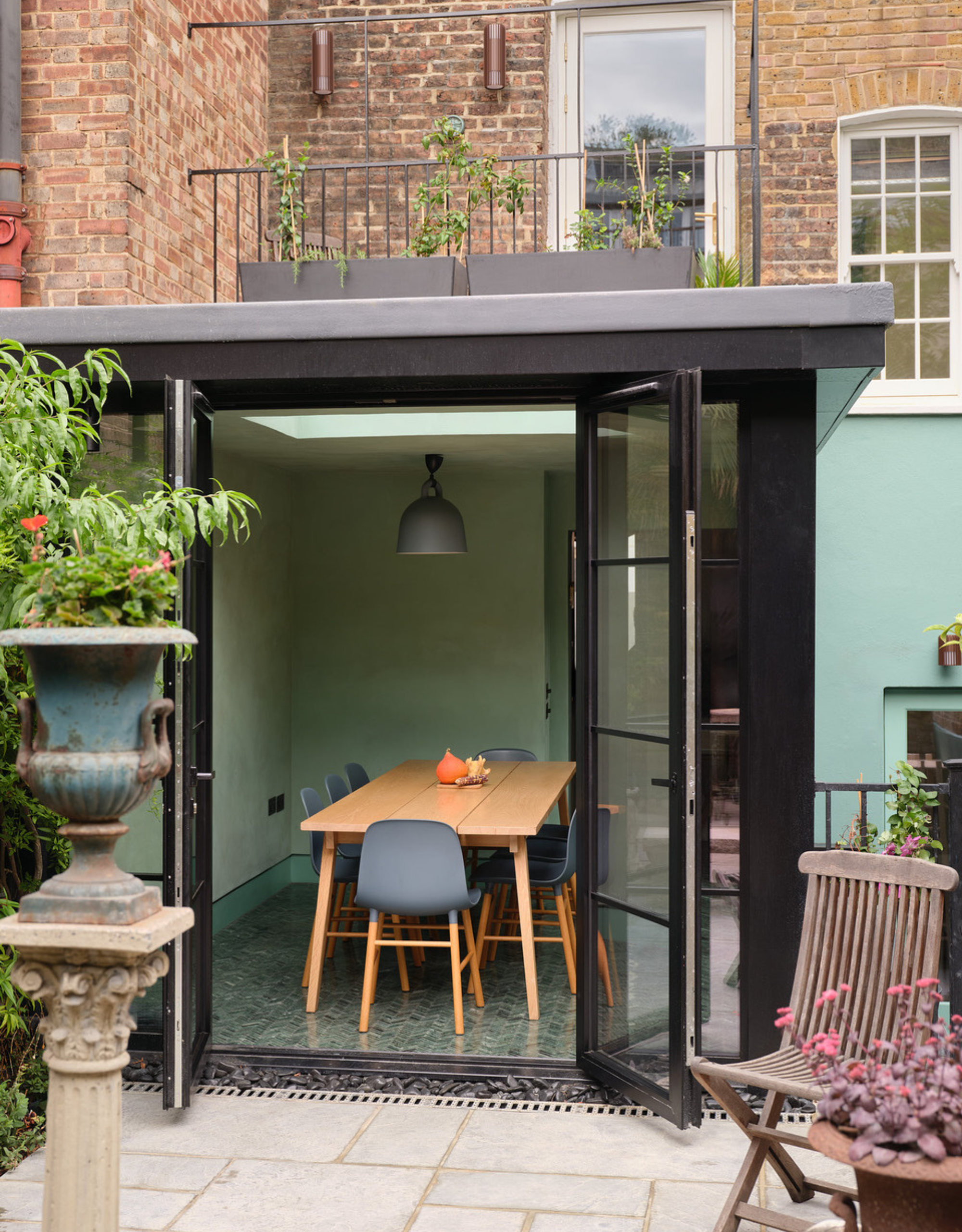 A small outdoor garden with a wooden chair, potted plants, and a patinaed planter on a plinth