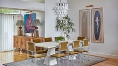 A white, modern dining space with a white table surrounded by mustard chairs in the centre of the space, with a grey rug underneath and white chandelier above.