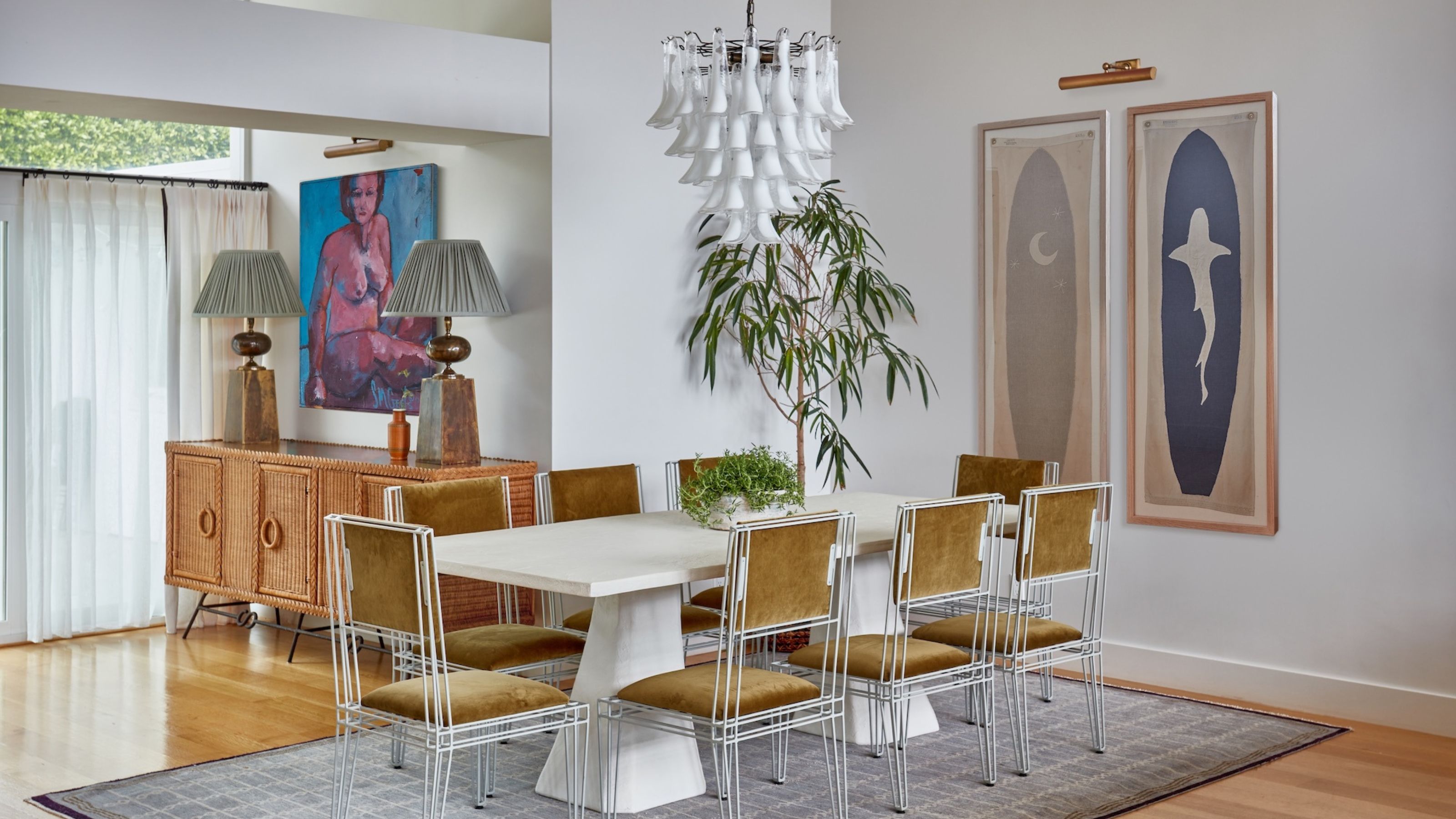 A white, modern dining space with a white table surrounded by mustard chairs in the centre of the space, with a grey rug underneath and white chandelier above.