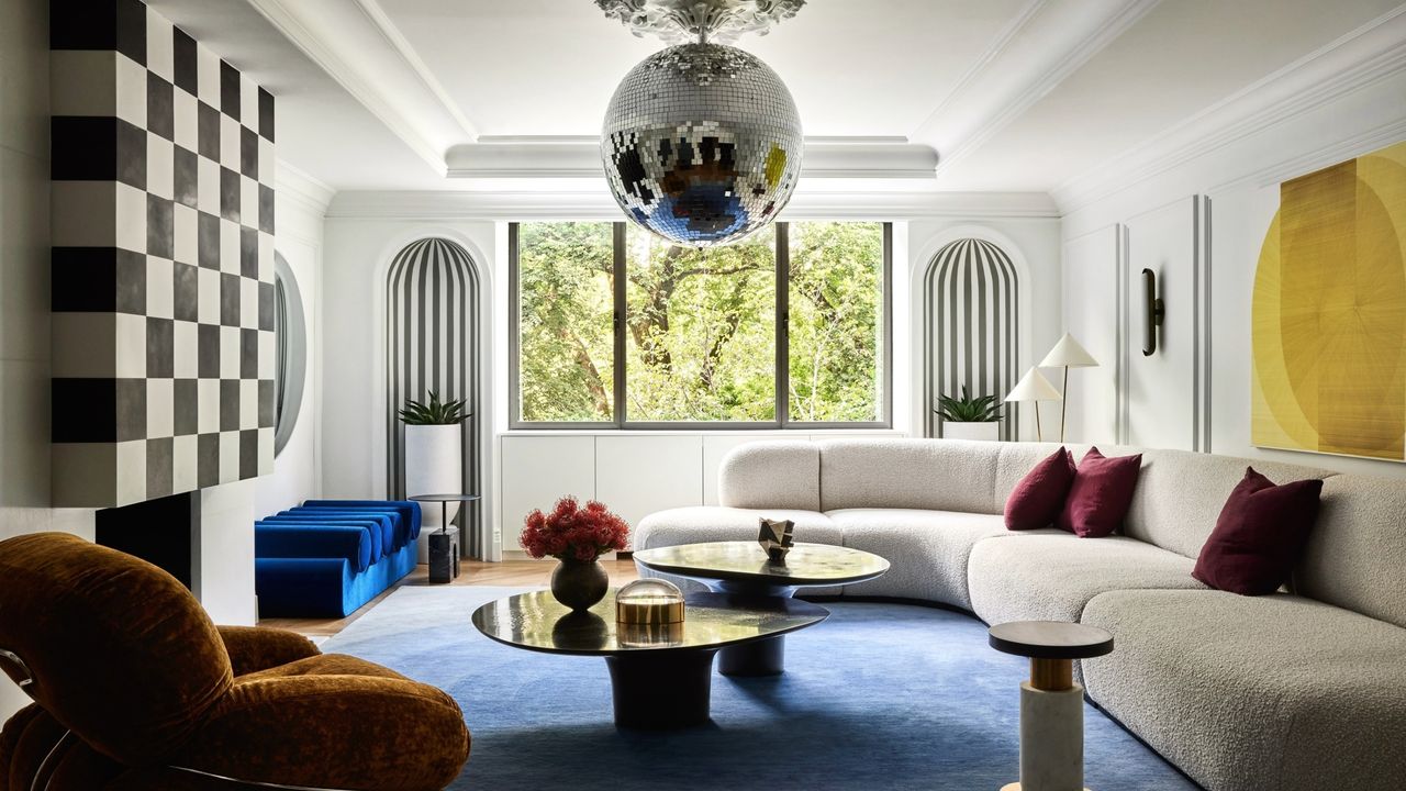 Image of a large, luxurious living room with white walls and a blue carpet. There is a long white curved sofa and nesting coffee tables. There is a disco ball hanging from the ceiling.