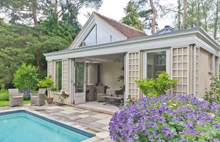 a chic and stylish grey/beige pool house with a domed roof, with grey garden furniture inside, plenty of hedges and lavender bushes outside, and a pool just in front