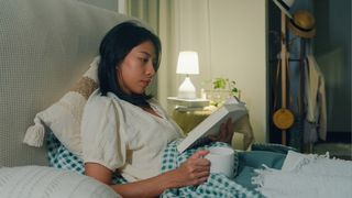 A woman in bed reading a book with a cup of tea in hand and a lamp on the bedside table, as she completes her nighttime routine