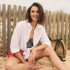 Model wearing a white shirt tied up with coral shorts and a wicker bag sold at Freemans sat on the beach
