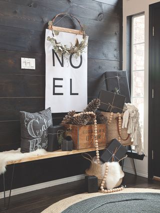 Christmas hallway/entry way with Noel hanging sign, bench with sheepskin, baskets and presents, Christmas cushion, box with huge pine cones, wooden beaded garland, rug