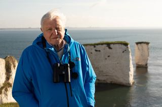 Sir David Attenborough stands on top of some cliffs, wearing a The North Face anorak and with binoculars around his neck. Behind him are the open sea and more cliffs.