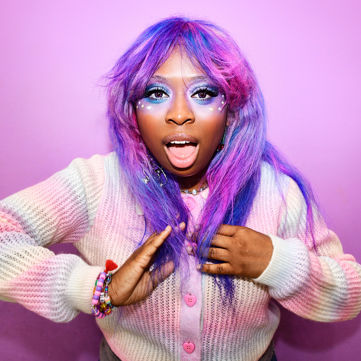 Photo of artist hemlocke springs standing in front of a purple backdrop. She is wearing a rainbow-colored cardigan with pink, heart-shaped buttons. Her hair is purple and pink. 