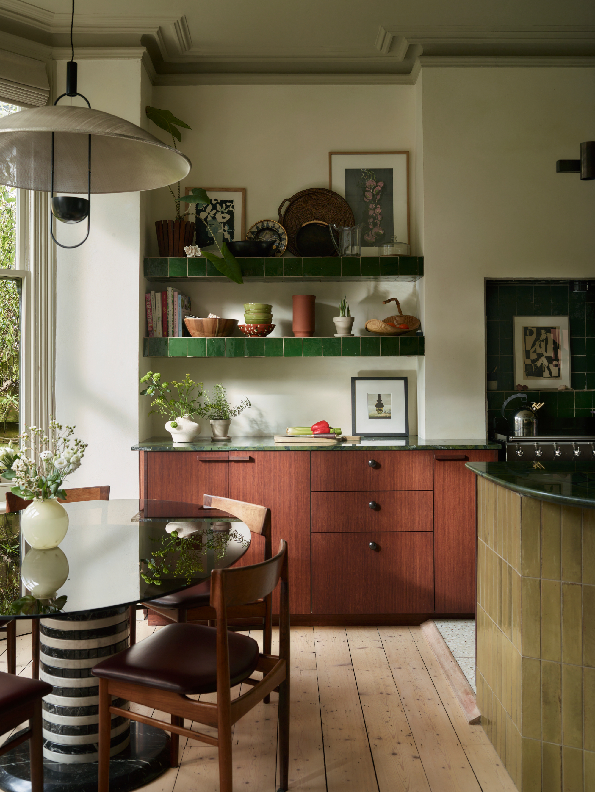 Corner of the kitchen with black and white striped breakfast table, dark wood chairs, red wood cabinets and green tile-lined shelving