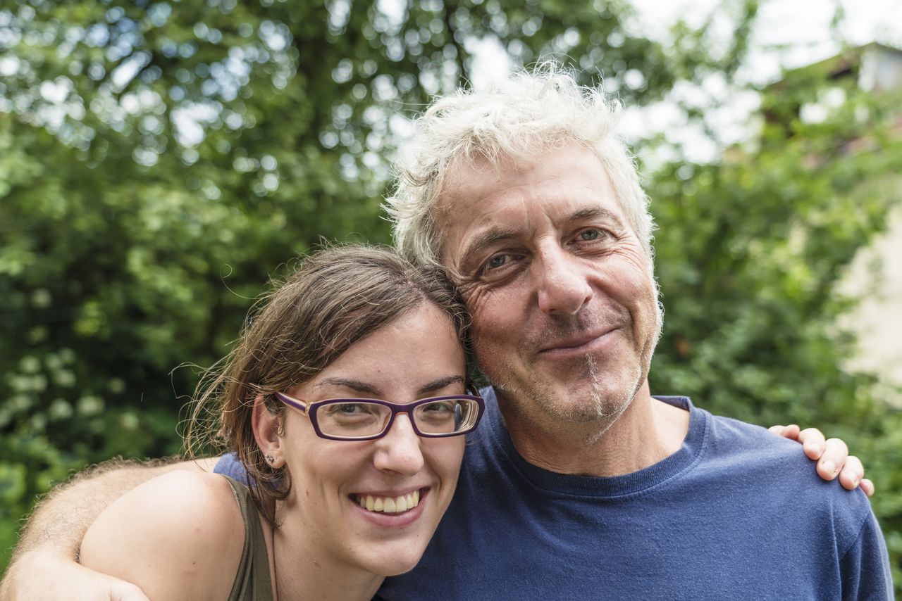 An older father and middle-aged daughter smile at the camera in their backyard.