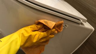 A microfiber cloth being used to clean the side of a toilet while wearing rubber gloves