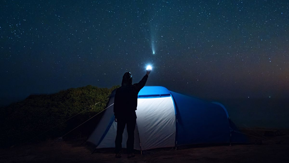 A man stands outside his tent in a quiet camping scene, pointing a flashlight directly at the comet Tsuchinshan-ATLAS (C/2023 A3), which shines among countless stars in the night sky, creating a captivating celestial moment.