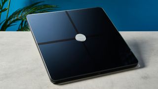 a modern black smart scale with a silver circle in its centre rests upon a light surface with a blue background