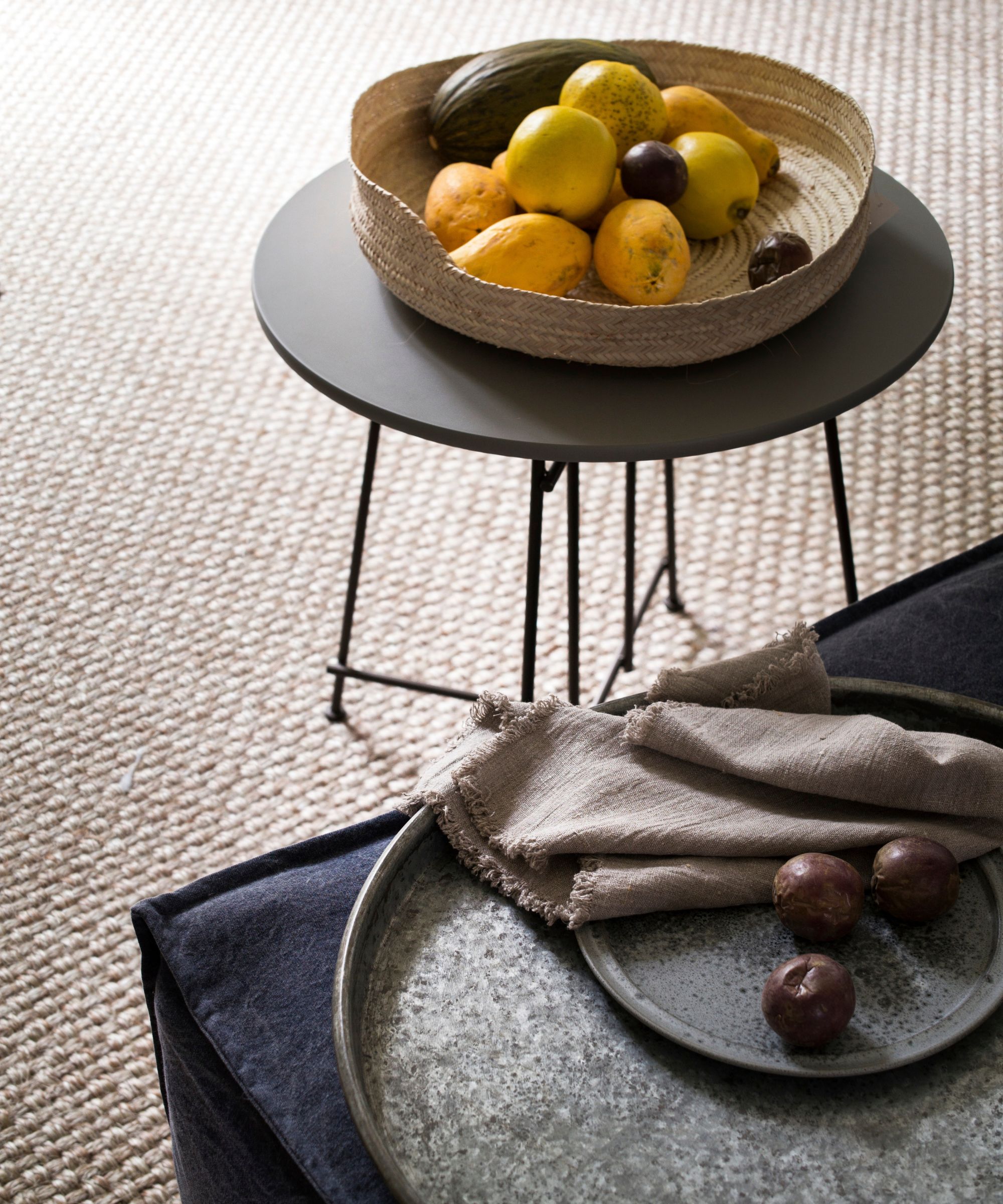 A rattan fruit bowl filled with fruit on a black circular table, with a dark blue sofa seen in the foreground with a silver metal tray, plate, three plums, and light brown napkin.