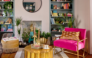 Room with two alcove bookshelves on either side of a fireplace, a pink armchair, a gold table and various plants