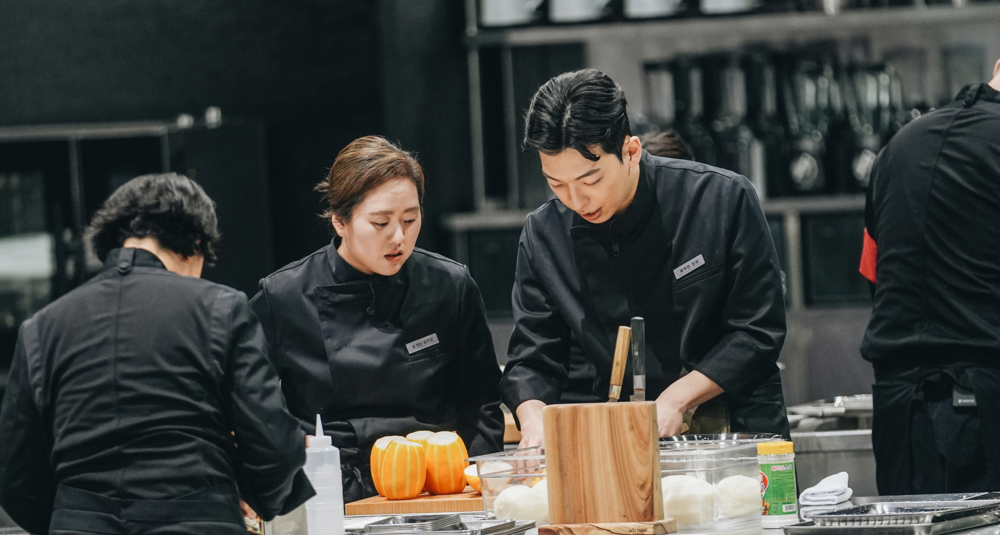 chefs in black coats preparing food over a silver counter top in an open kitchen in a still from the netflix korean reality show culinary class wars season 2