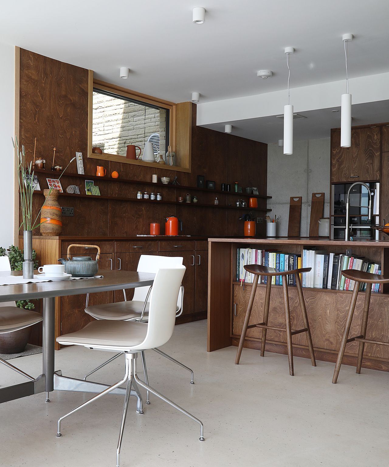 A dark wood kitchen with island and open plan dining area. There are stools pushed up to the kitchen island which has cupboards and shelving built in