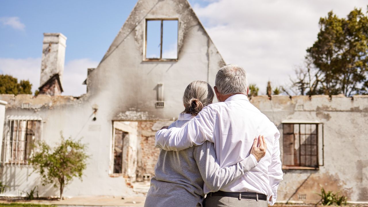 A senior couple hugging and comforting each other while staring at the home they have lost to a fire