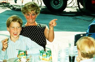 Princess Diana wearing a black polka dot dress and pointing as Prince William eats a snack and laughs at a picnic table