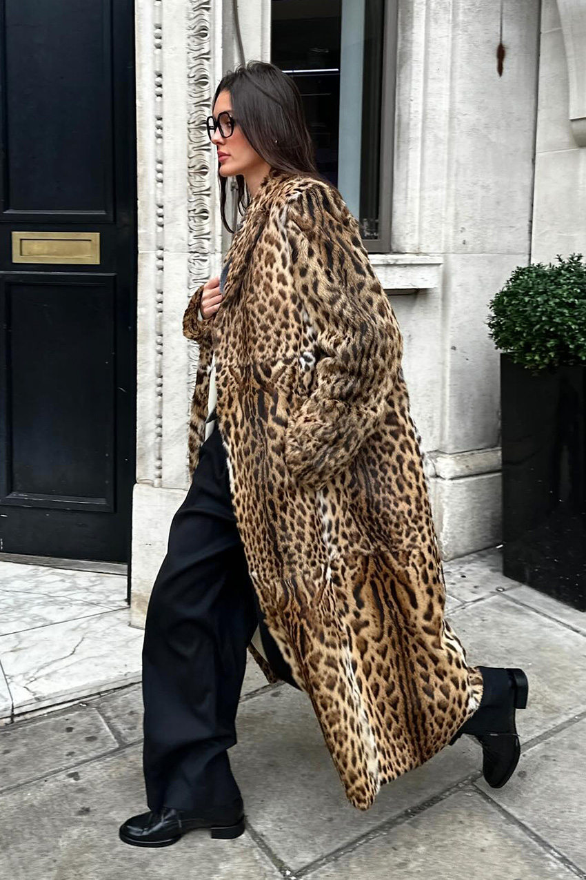 The best statement coats are shown in a street style photo of a woman standing on the sidewalk wearing oversized black eyeglasses with a long leopard print fur coat styled over a white-shirt, gray cardigan, black trousers, and black ankle boots