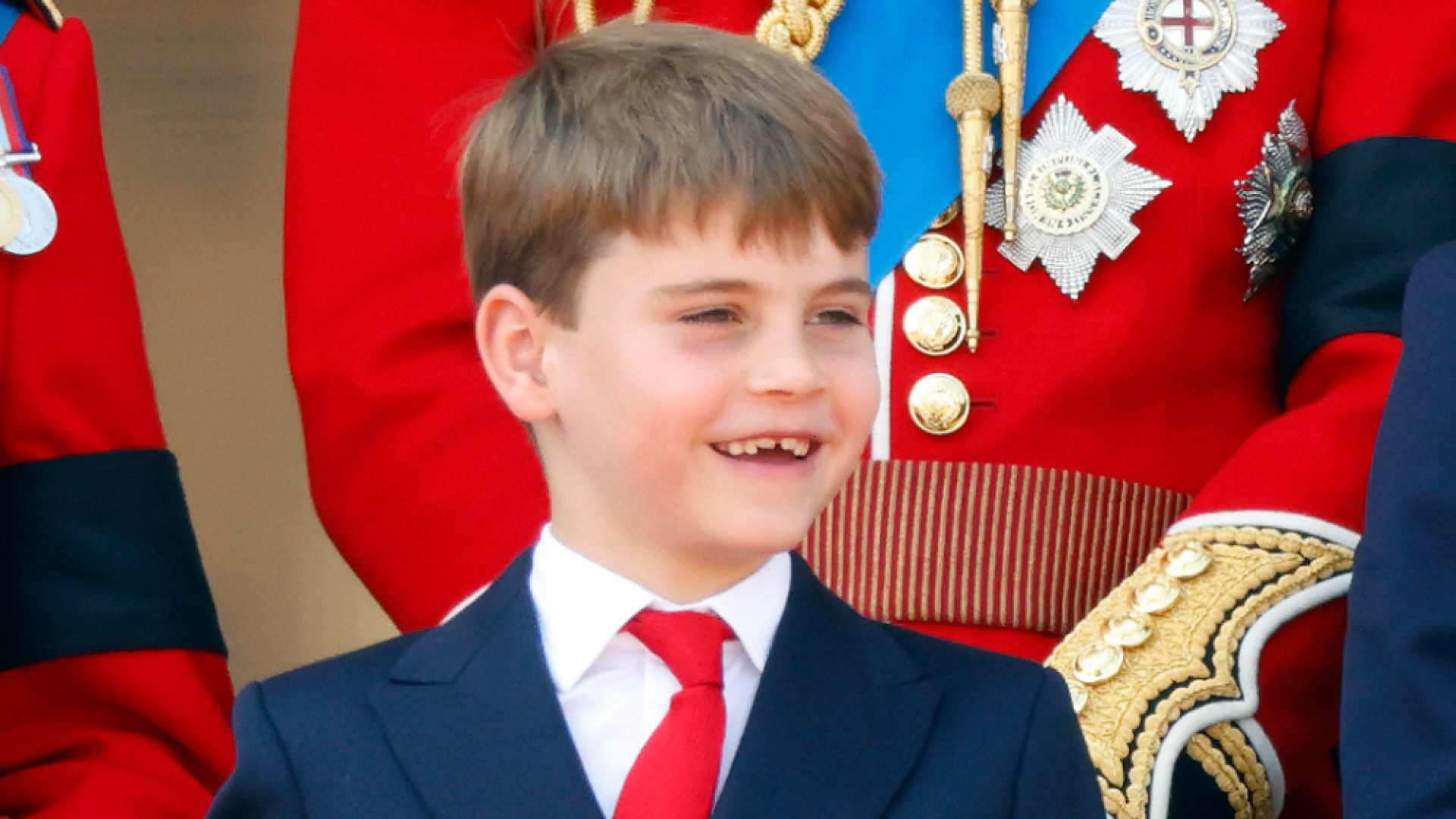 Prince Louis smiles, showing off his growing teeth, while wearing a navy suit with a white shirt and red tie