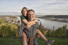A man in his 50s gives a piggyback ride to his female partner. They are hiking and there's a view of a river behind them. They both look very happy.