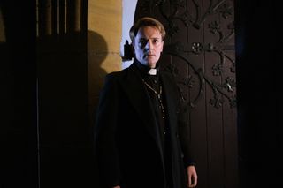 Father Michael (Joe Barnes) stands in near darkness outside the door of the chapel with a very serious look on his face