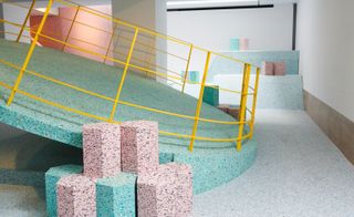 RIBA’s The Brutalist Playground, pastel green and pink speckled blocks at the base of a metal yellow framed circular tilted green speckled platform, grey speckled walkway and white walls with brown base trim