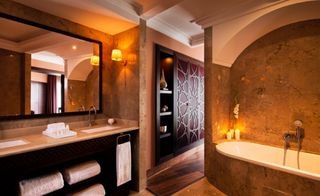 A hotel bathroom with a white tub, long counter, twin sinks, twin mirrors, wooden shelves and brown marble walls.