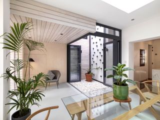 A sitting area with a glass table, wooden chairs, potted plants, a skylight and light tiles flooring and walls.