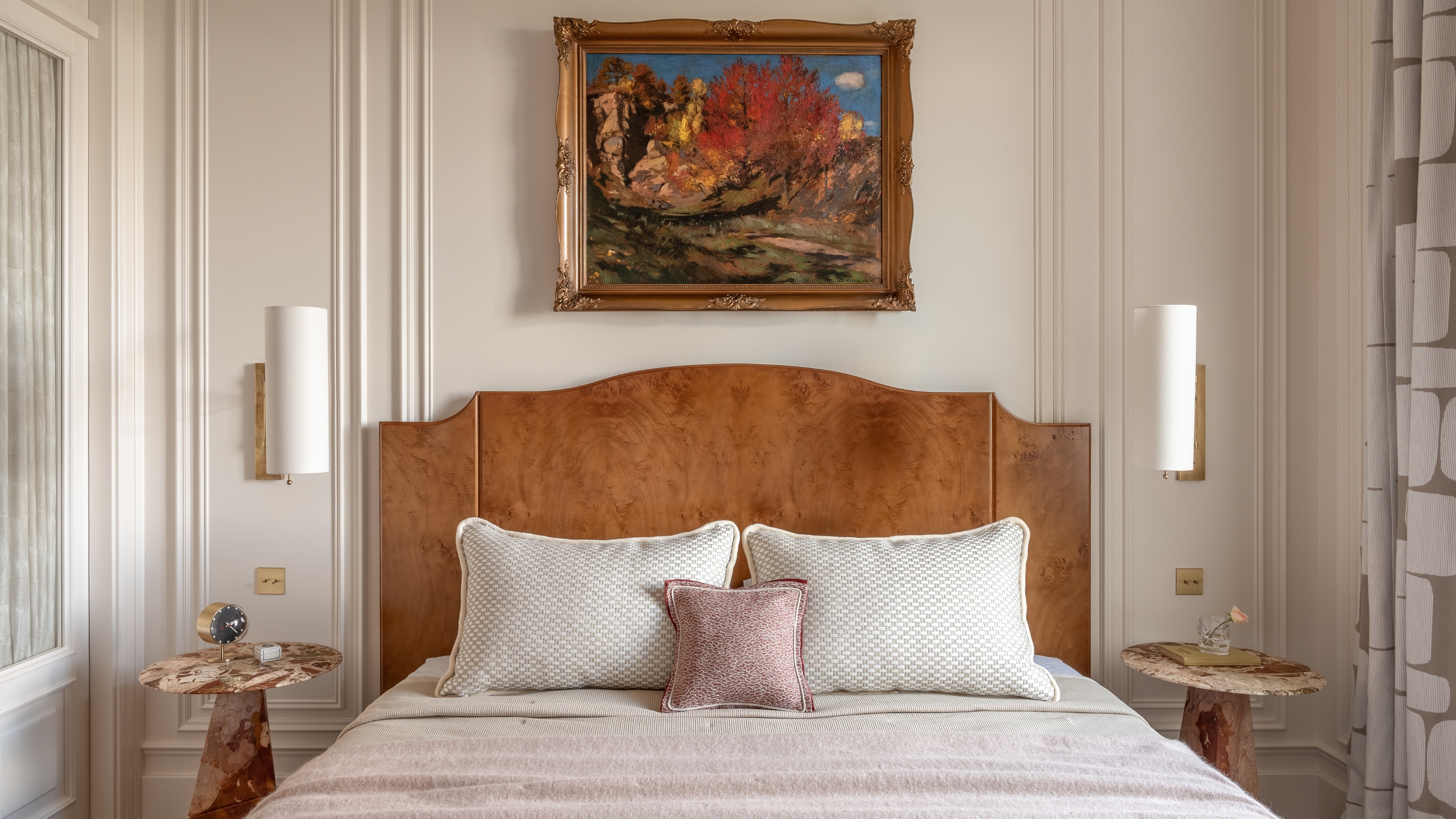 Image of a beige bedroom with a bed that has a wooden headboard and beige and rose pink bedding. There are marble side tables on either side of the bed with sconce lighting hanging above them.